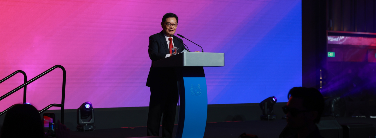 Man in suit speaks at podium with SG coat of arms, pink/blue backdrop. Lights and partial audience visible.
